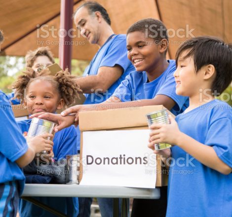 Multi-ethnic group of children on a soccer team work together to organize a charity drive for a local natural disaster event in their area.  The children set up a table at their local park to gather canned goods and clothing for the victims affected by the disaster.  Their team coach helps with the event.